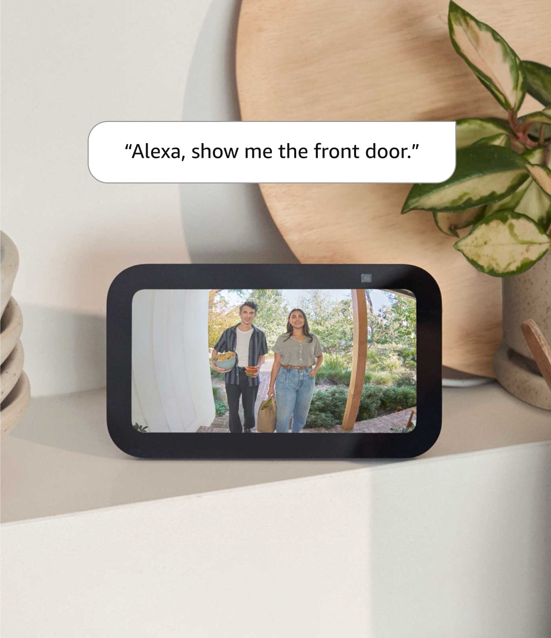 Echo Show 5: 147.3mm x 86.4mm (5.8 by 3.4 inch) horizontal tablet sitting on a shelf. Screen shows a man and woman on a front porch holding snacks. Text reads: “Alexa, show me the front door.”