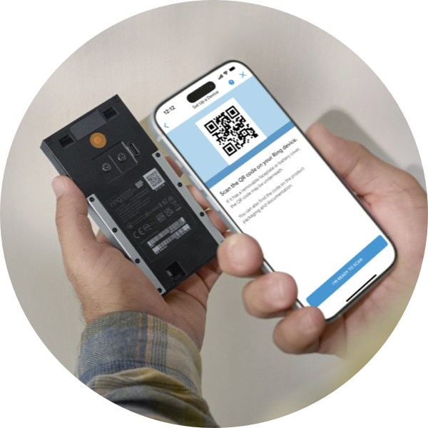 A pair of hands holding a phone and Ring Battery Doorbell. The phone screen shows a QR code, instructing the user to scan a QR code on the back of their device.
