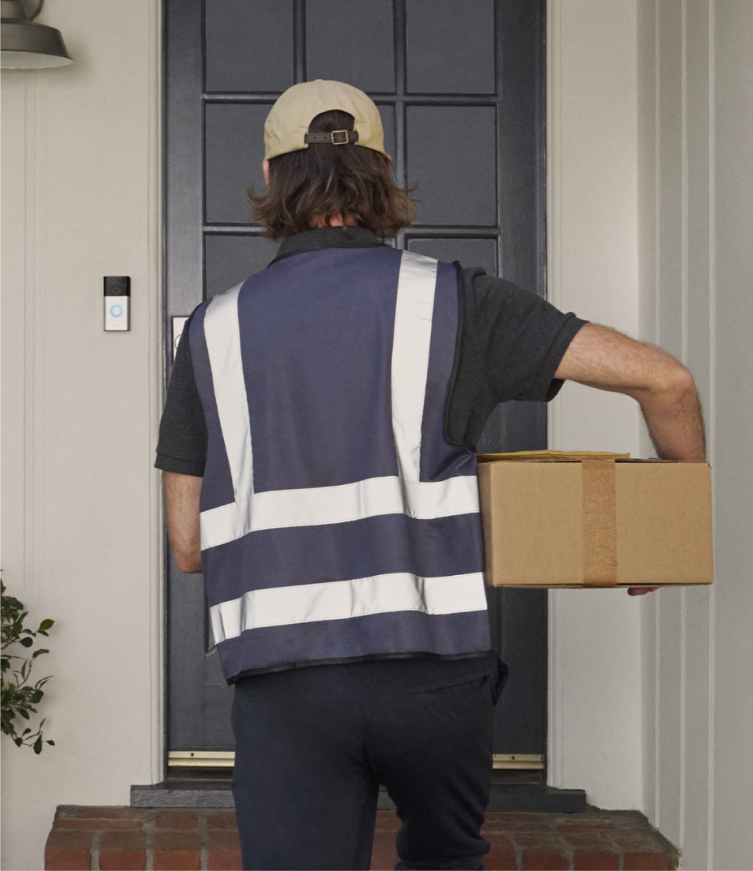 Ring Battery Doorbell: 4.9 by 2.4 inch video doorbell on a wall next to a delivery man facing the front door while holding a box.